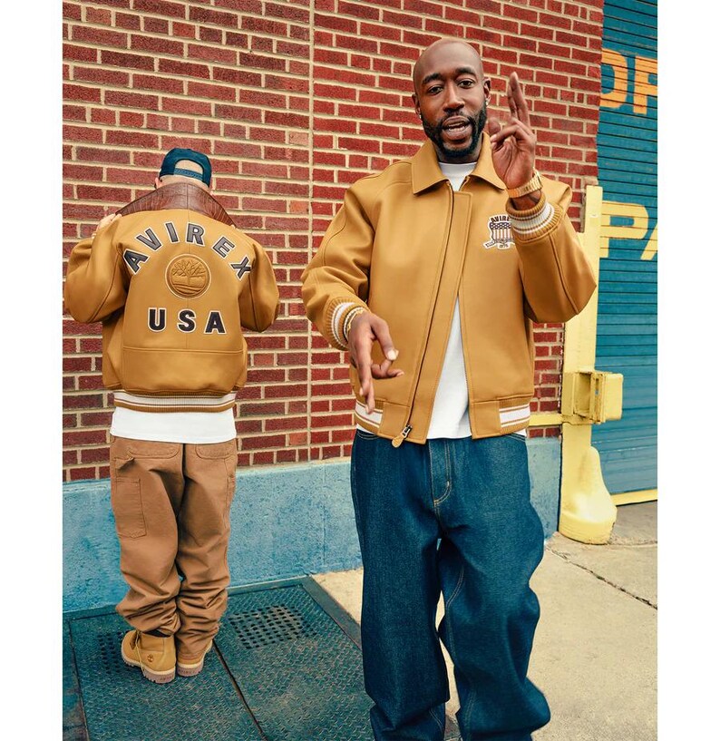 May include: Two people wearing mustard-coloured jackets with white and brown accents. One jacket has "AVIREX USA" on the back. One person wears blue jeans, the other wears brown trousers. The background is a brick wall.