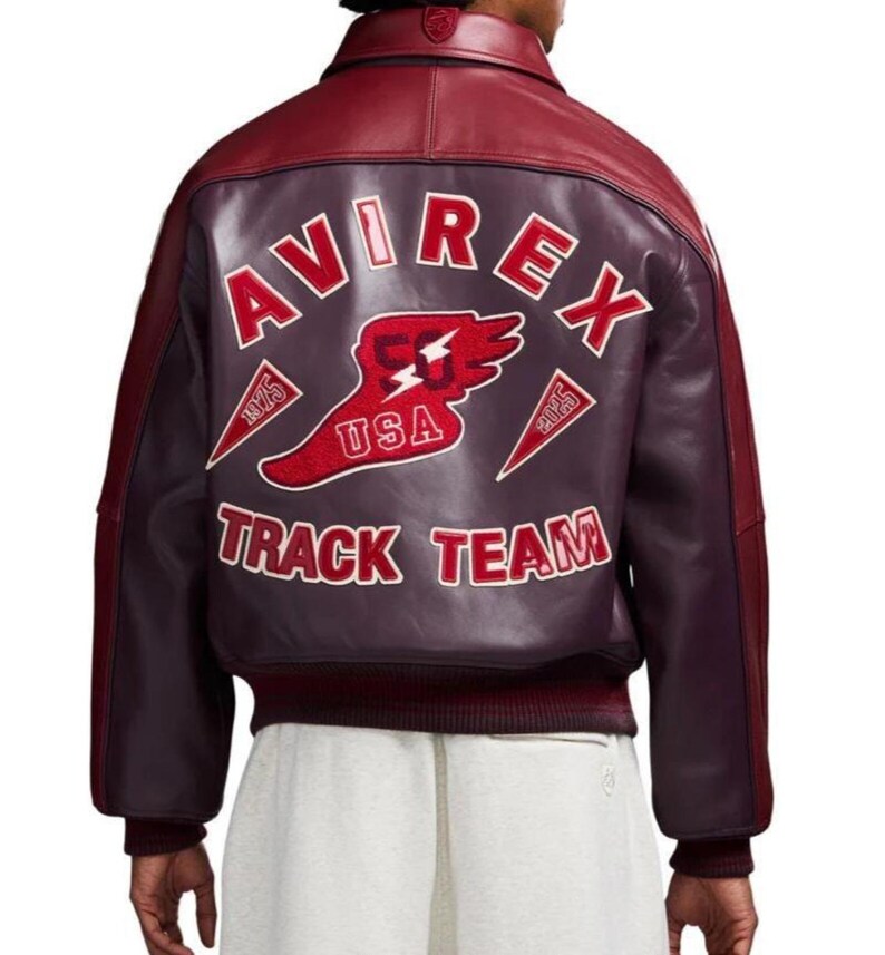 Puede incluir: Chaqueta de cuero burdeos con cuello y mangas en rojo oscuro en contraste. La espalda presenta el texto "AVIREX TRACK TEAM" en blanco, con un emblema de EE. UU. rojo y blanco. La chaqueta tiene dobladillo y pu&ntilde;os acanalados.