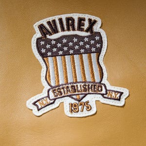 May include: Embroidered patch featuring the text "AVIREX", a shield design with stars and stripes, and the words "ESTABLISHED NY 1975". The patch is white, brown, gold, and blue, set against a tan backdrop.