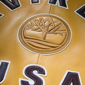 May include: Close-up of a mustard-coloured leather jacket with a circular embroidered patch featuring a tree design. The patch is surrounded by a white border and brown stitching. Brown and white letter patches are also visible.