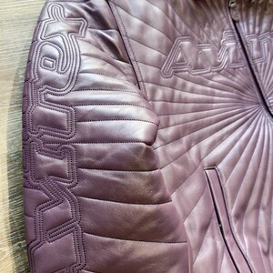 May include: A mauve leather jacket with quilted diamond pattern and the word "AMU" stitched across the chest. The left sleeve features a stylized design. The jacket has a collared neckline and a zippered front.