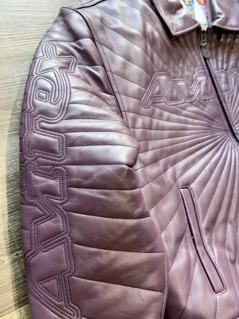 May include: A mauve leather jacket with quilted diamond pattern and the word "AMU" stitched across the chest. The left sleeve features a stylized design. The jacket has a collared neckline and a zippered front.