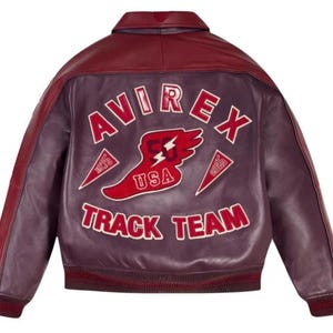 Puede incluir: Chaqueta de cuero burdeos y granate con el texto "AVIREX TRACK TEAM" en letras blancas. La parte trasera de la chaqueta presenta un gr&aacute;fico rojo y blanco con las palabras "USA" y "53". La chaqueta tiene cuello y pu&ntilde;os acanalados.