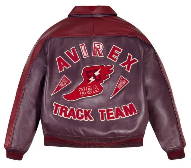 Puede incluir: Chaqueta de cuero burdeos y granate con el texto "AVIREX TRACK TEAM" en letras blancas. La parte trasera de la chaqueta presenta un gr&aacute;fico rojo y blanco con las palabras "USA" y "53". La chaqueta tiene cuello y pu&ntilde;os acanalados.