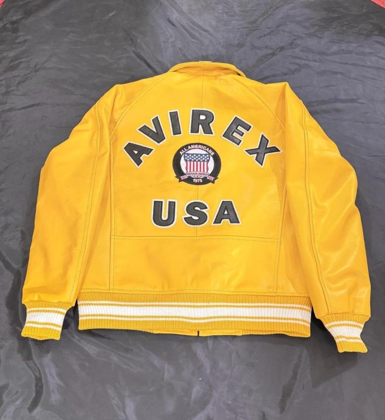 May include: A bright yellow leather jacket featuring the word "AVIREX" in black lettering across the back. Below the word "AVIREX" is a circular patch with the words "ALL AMERICANS 1975" and a design of the American flag. The word "USA" is below the patch.