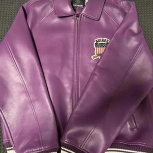 Puede incluir: Chaqueta de cuero morada con cremallera y cuello. La chaqueta presenta un parche en el pecho con el texto "AVIREX" y un dise&ntilde;o de bandera estadounidense. Los pu&ntilde;os y el dobladillo son acanalados con rayas blancas.