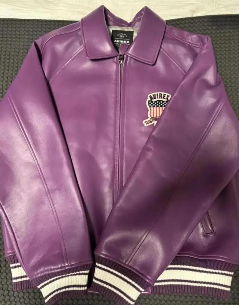 Puede incluir: Chaqueta de cuero morada con cremallera y cuello. La chaqueta presenta un parche en el pecho con el texto "AVIREX" y un dise&ntilde;o de bandera estadounidense. Los pu&ntilde;os y el dobladillo son acanalados con rayas blancas.
