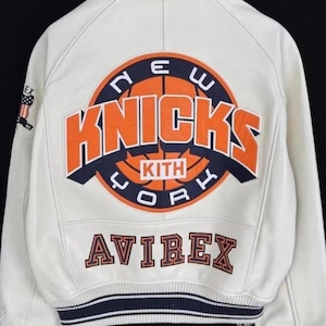 Pode incluir: Casaco de couro creme com o logotipo do New York Knicks em laranja e azul marinho nas costas. O casaco inclui a palavra "AVIREX" em letras laranja e uma faixa listrada azul marinho e branca.