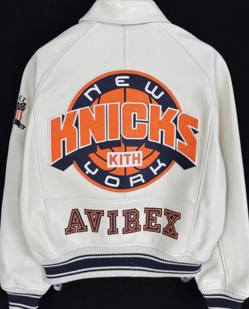 Pode incluir: Casaco de couro creme com o logotipo do New York Knicks em laranja e azul marinho nas costas. O casaco inclui a palavra "AVIREX" em letras laranja e uma faixa listrada azul marinho e branca.