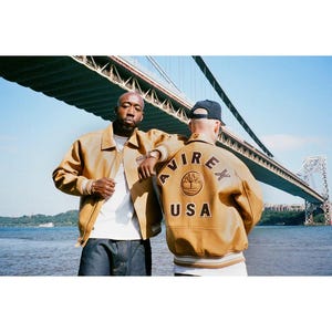 May include: Two people wearing tan leather jackets with the text "AVIREX USA" on the back. The jackets have a ribbed collar and cuffs. The background features a bridge over a body of water.