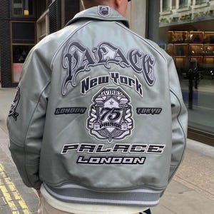 May include: A light grey leather jacket with the word "PALACE" in large, stylized lettering across the back, along with "New York", "London", and "Tokyo". The jacket features a patch commemorating "75 Palace Pride" and the Avirex brand.
