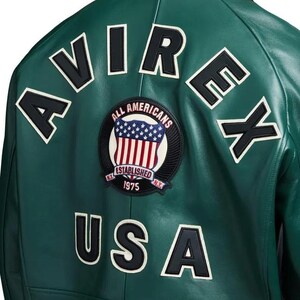 Może przedstawiać: Ciemnozielona sk&oacute;rzana kurtka z czarnymi literami obrysowanymi na biało, kt&oacute;re piszą "AVIREX" i "USA". Okrągła naszywka przedstawia tarczę z amerykańską flagą i napisami "ALL AMERICANS ESTABLISHED 1975 N.Y."