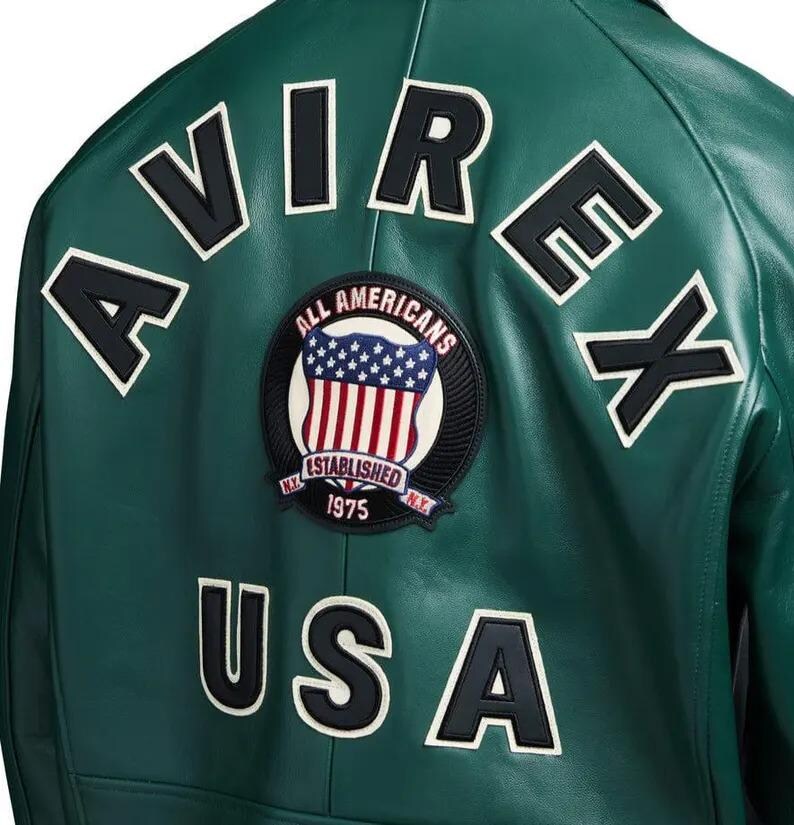 Może przedstawiać: Ciemnozielona sk&oacute;rzana kurtka z czarnymi literami obrysowanymi na biało, kt&oacute;re piszą "AVIREX" i "USA". Okrągła naszywka przedstawia tarczę z amerykańską flagą i napisami "ALL AMERICANS ESTABLISHED 1975 N.Y."