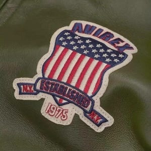 May include: Close-up of a green leather jacket with a patch. The patch features the word "AVIREX" in blue, a shield with red and white stripes and blue stars, and the text "N.Y. ESTABLISHED 1975 N.Y." in a banner.