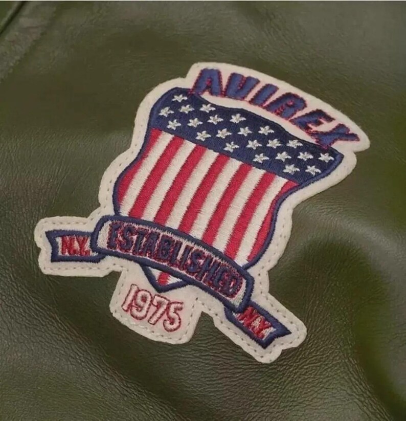 May include: Close-up of a green leather jacket with a patch. The patch features the word "AVIREX" in blue, a shield with red and white stripes and blue stars, and the text "N.Y. ESTABLISHED 1975 N.Y." in a banner.