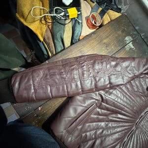 May include: A maroon leather jacket with quilted stitching and a ribbed cuff. The jacket features the word "AMU" stitched on the sleeve. The jacket is on a wooden surface.