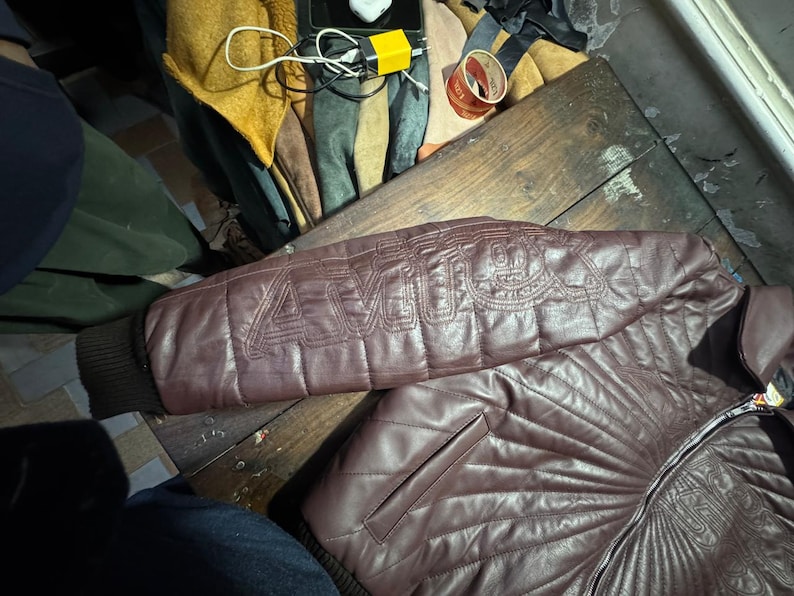 May include: A maroon leather jacket with quilted stitching and a ribbed cuff. The jacket features the word "AMU" stitched on the sleeve. The jacket is on a wooden surface.