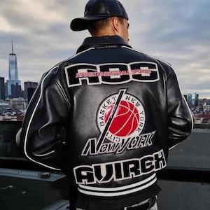 Peut inclure: Une veste universitaire en cuir noir avec des passepoils blancs et de grands &eacute;cussons brod&eacute;s au dos. Le texte indique "AOS AVIREX BASKETBALL SPORTS" et "NEW YORK" avec un motif central de ballon de basket rouge et "AVIREX" en bas.