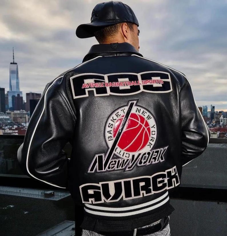 Peut inclure: Une veste universitaire en cuir noir avec des passepoils blancs et de grands &eacute;cussons brod&eacute;s au dos. Le texte indique "AOS AVIREX BASKETBALL SPORTS" et "NEW YORK" avec un motif central de ballon de basket rouge et "AVIREX" en bas.