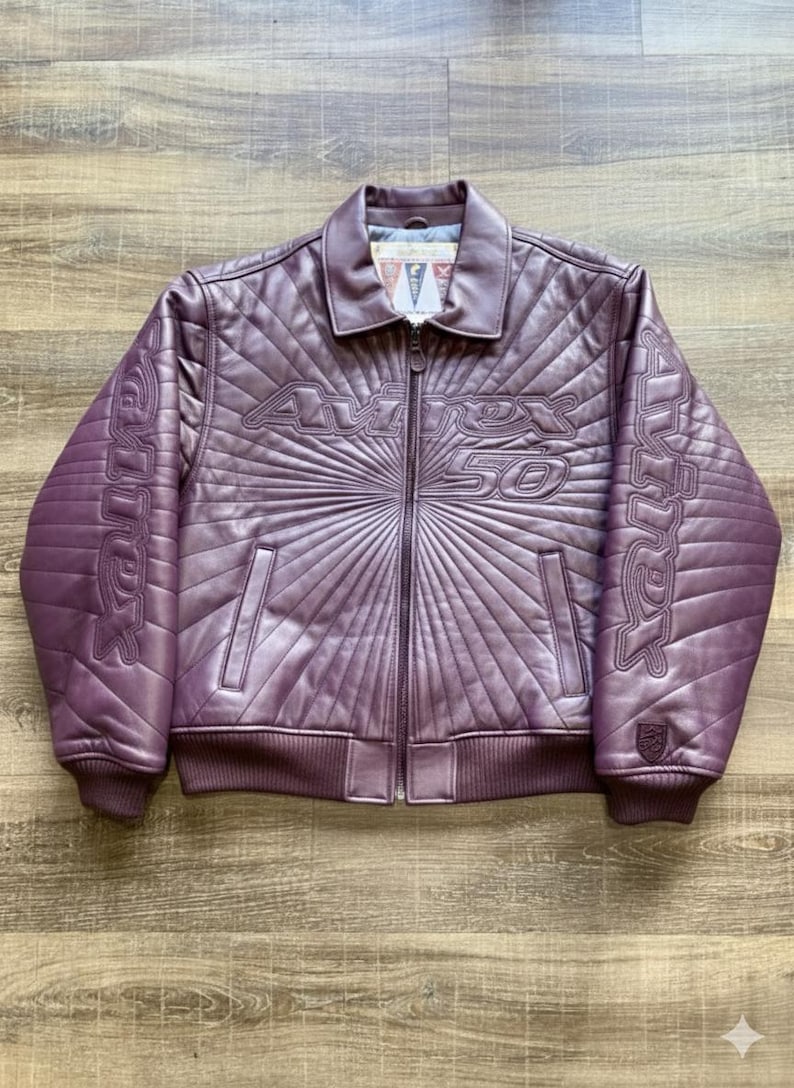 May include: A purple leather jacket with a quilted design and a zip-up front. The jacket features the text "AVIREX 50" across the chest and on the sleeves. It has a collared neckline and ribbed cuffs and hem.