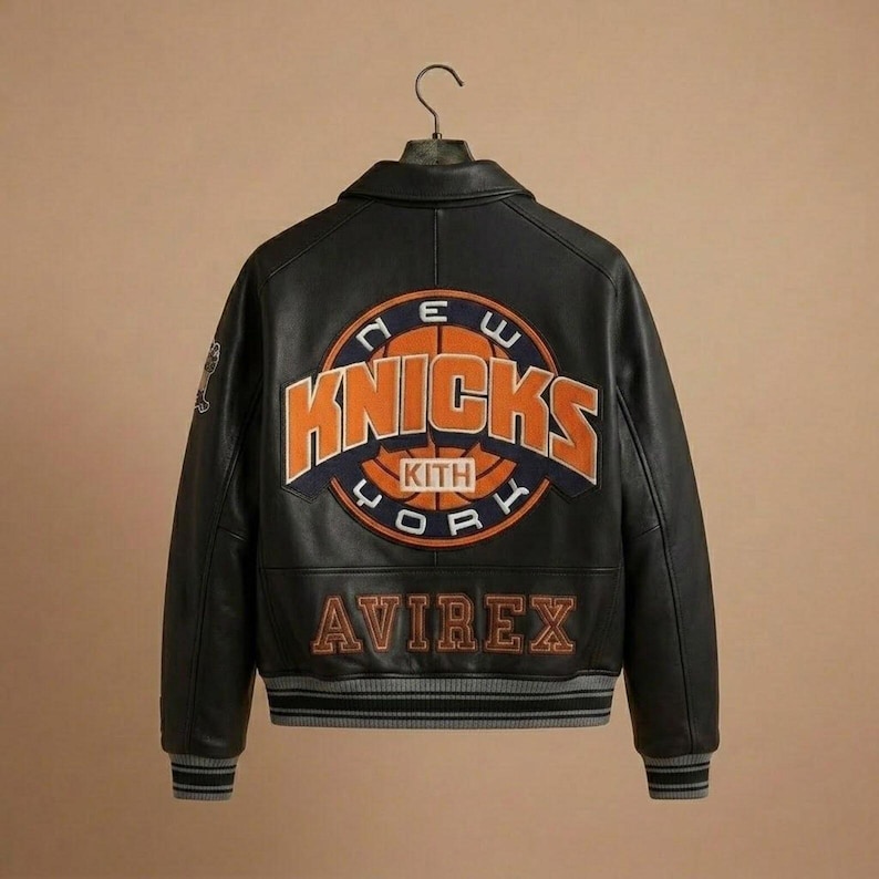 Puede incluir: Una chaqueta universitaria de cuero negro con un gran logotipo de baloncesto de los New York Knicks bordado en la espalda, que incluye el texto KITH en el centro. La palabra AVIREX est&aacute; bordada debajo en naranja y tiene pu&ntilde;os acanalados a rayas.