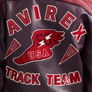 Puede incluir: Primer plano de una chaqueta de cuero burdeos con parches bordados en rojo y blanco. Los parches incluyen la palabra "AVIREX", "TRACK TEAM", "USA" y los n&uacute;meros "1975" y "2025". La chaqueta tiene un cuello rojo.