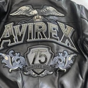 May include: Black leather jacket with embroidered patches. The back features the word "AVIREX" in large, outlined letters, an eagle with spread wings, and a shield with the number "75" and the text "AVIATOR'S KING".