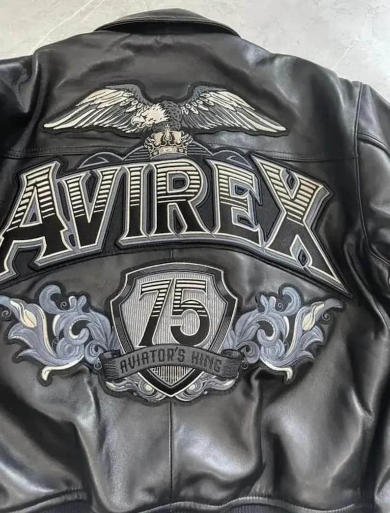 May include: Black leather jacket with embroidered patches. The back features the word "AVIREX" in large, outlined letters, an eagle with spread wings, and a shield with the number "75" and the text "AVIATOR'S KING".