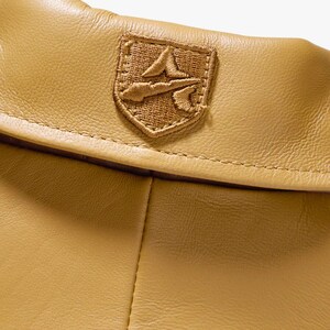 May include: Close-up of a tan leather item with a stitched seam. A brown embroidered emblem is sewn onto the leather. The emblem features a stylized design within a shield shape.