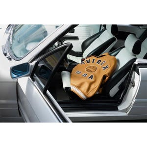 May include: A tan leather jacket with the text "AVIREX USA" is inside a white and black car. The jacket has white stripes on the sleeves and collar. The car's interior is white and black.