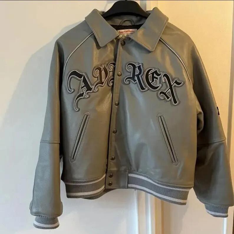 May include: Gray leather jacket with the word "AVIREX" in black script across the chest. The jacket has a collared neckline, snap closures, and striped cuffs and hem. The jacket is hanging on a black hanger.