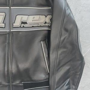 May include: Black leather jacket featuring the word "REX" in a grey and white design. The jacket has a zip closure, ribbed hem, and cuffs with grey and white stripes. The text "NITORUN" is below the word "REX".
