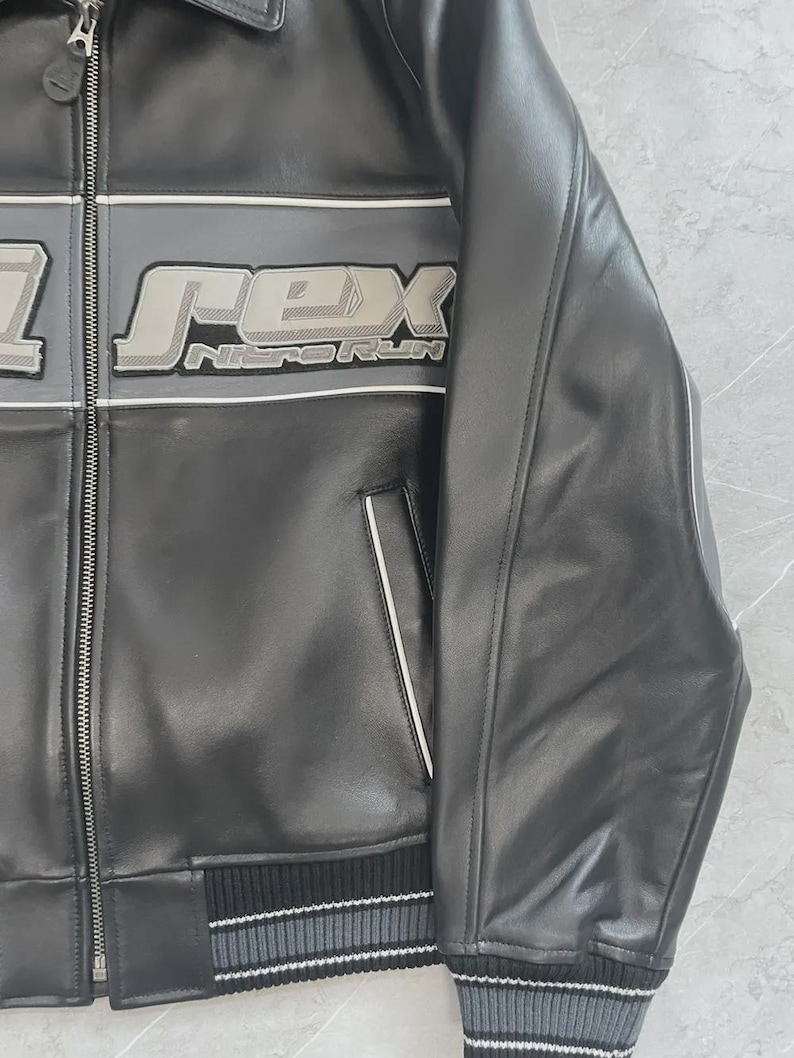 May include: Black leather jacket featuring the word "REX" in a grey and white design. The jacket has a zip closure, ribbed hem, and cuffs with grey and white stripes. The text "NITORUN" is below the word "REX".