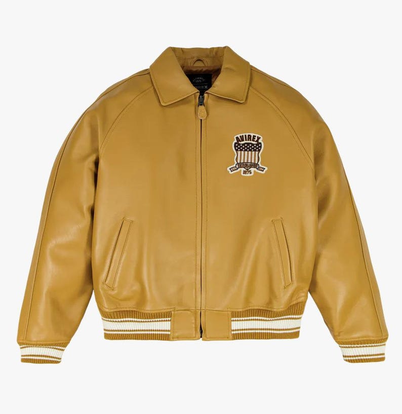 May include: Mustard yellow leather bomber jacket with a zip-up front, side pockets, and a ribbed hem and cuffs with white stripes. Features a patch on the chest with the text "AVIREX" and a patriotic design.