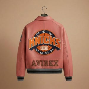 Puede incluir: Una chaqueta universitaria de cuero rosa polvoriento con un gran parche circular en la espalda con el texto NEW YORK KNICKS KITH en naranja y azul marino. La palabra AVIREX est&aacute; bordada en letras grandes de bloque debajo del parche. La chaqueta tiene cuello, mangas largas y pu&ntilde;os y dobladillo acanalados a rayas.