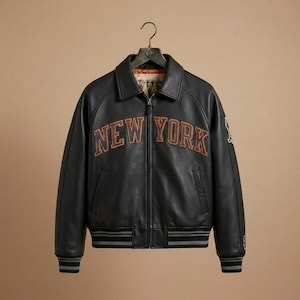 Puede incluir: Una chaqueta de cuero negra con cuello cl&aacute;sico, cremallera frontal completa y pu&ntilde;os y dobladillo acanalados con rayas grises. El texto NEW YORK est&aacute; bordado en el pecho con letras naranjas y negras, con parches adicionales en la manga.