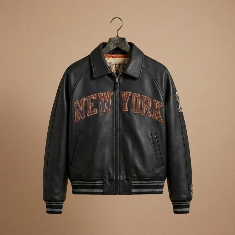 Puede incluir: Una chaqueta de cuero negra con cuello cl&aacute;sico, cremallera frontal completa y pu&ntilde;os y dobladillo acanalados con rayas grises. El texto NEW YORK est&aacute; bordado en el pecho con letras naranjas y negras, con parches adicionales en la manga.