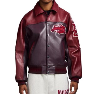 Puede incluir: Chaqueta de cuero burdeos y burdeos oscuro con cierre de botones, cuello y pu&ntilde;os y dobladillo acanalados. La chaqueta presenta un parche con el texto "AVIREX" y el n&uacute;mero "74" en la manga.