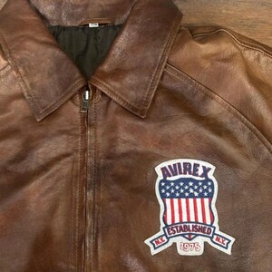 May include: Brown leather jacket with a zip-up front and a collared neckline. The jacket features a patch on the chest with the text "AVIREX ESTABLISHED N.Y. 1975" and a design of the American flag.