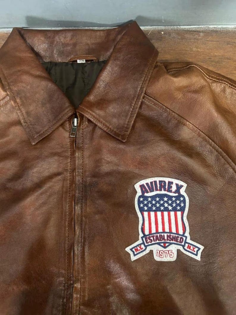 May include: Brown leather jacket with a zip-up front and a collared neckline. The jacket features a patch on the chest with the text "AVIREX ESTABLISHED N.Y. 1975" and a design of the American flag.