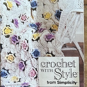 May include: A vintage Simplicity crochet pattern booklet titled 0423 Afghans to Crochet. The cover features a white crocheted afghan decorated with three-dimensional pink, yellow, and blue flowers, draped over a white wicker chair.