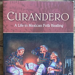 May include: A paperback book titled Curandero: A Life in Mexican Folk Healing by Eliseo Cheo Torres with Timothy L. Sawyer, Jr. The cover features a painting of three people in a dimly lit room with a candle, colorful patterned clothing, and tools.