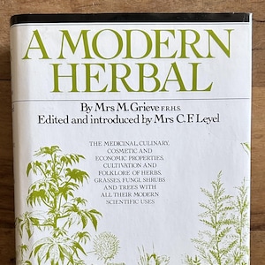 May include: A white book cover titled A MODERN HERBAL by Mrs M. Grieve, edited by Mrs C. F. Leyel. The cover features green botanical illustrations of various plants, herbs, and shrubs, along with descriptive text about medicinal and culinary uses.