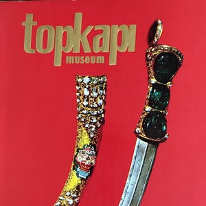 May include: A red book cover titled Topkapi Museum featuring a gold-toned dagger with three large green stones on the hilt and a matching curved scabbard decorated with small white stones and a floral motif, with text listing the contents below.