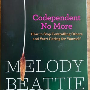May include: A book titled "Codependent No More" by Melody Beattie. The cover is dark green with white text and a dandelion illustration. A silver sticker indicates it's the 25th Anniversary Edition, with over 5 million copies sold.