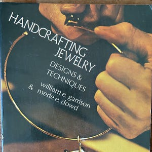 May include: A book titled "Handcrafting Jewelry: Designs & Techniques" featuring a gold necklace on the cover. The cover art includes a close-up of a person working on jewelry. The book's authors are William E. Garrison and Merle E. Dowd.