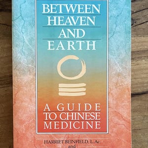 May include: A paperback book titled Between Heaven and Earth: A Guide to Chinese Medicine by Harriet Beinfield and Efrem Korngold. The cover features a gradient from blue to orange with a central circular symbol and white text throughout.
