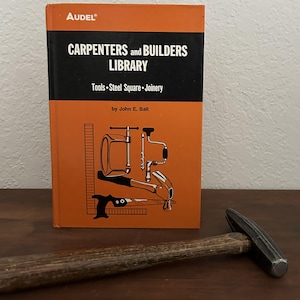 May include: An orange and black book titled "Carpenters and Builders Library" with illustrations of tools. A hammer with a wooden handle rests in front of the book, suggesting a carpentry theme.