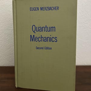 May include: A hardcover textbook titled Quantum Mechanics, Second Edition by Eugen Merzbacher. The book features a light olive green cover with dark blue text printed on the front, displaying the author's name, the title, and the edition.