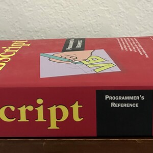 May include: A red VBScript Programmer's Reference book with yellow text on the cover. The cover features a graphic of a hand holding a green pen over a grid with the letters VB. The Wrox logo is visible on the side and the spine of the book.